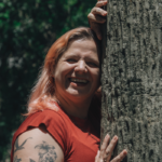 Joyful woman with tattoos hugging a tree in a sunny park setting.