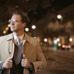 Confident adult male in formal wear standing on night street and holding collars of trench while looking away